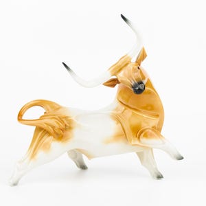 May include: A ceramic bull figurine featuring a glossy finish with a white base and tan-coloured patches. The bull is depicted in a dynamic pose with long, curved white horns that have black tips, a raised tail, and a dark muzzle and nose detail.