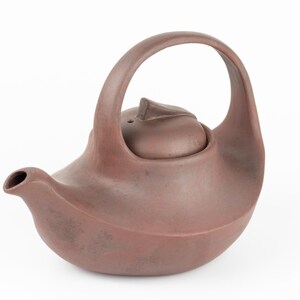 以下が含まれることがあります： 曲線的なハンドルと蓋が付いた茶色のセラミックティーポット。ティーポットには注ぎ口と丸みを帯びたボディがあります。ハンドルはアーチ型で、蓋には小さなノブが付いています。ティーポットは赤茶色で、白い背景に置かれています。