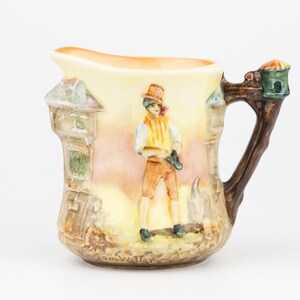 May include: A vintage ceramic jug featuring a painted scene of a man in a hat and shorts. The handle is designed to look like a tree branch. The jug has a light yellow and brown colour scheme.