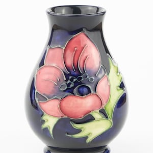 Könnte beinhalten: Eine dunkelblaue Keramikvase mit floralem Design. Die Vase zeigt eine große rosa und rote Blume mit grünen Blättern. Die Blume ist der Mittelpunkt, vor dem dunkelblauen Hintergrund. Die Vase hat eine abgerundete Form und einen kleinen Sockel.