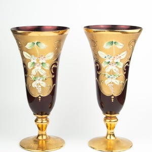 May include: Two ornate, stemmed glasses. The upper portion is gold with floral designs and a deep burgundy base. The rim is a deep burgundy color. The base and stem are gold. The glasses are likely for decorative use.
