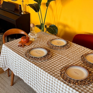 May include: A brown and white gingham tablecloth with four place settings. Each place setting includes a woven placemat, a round white plate with a French phrase, and a silver fork. The phrases on the plates are "Bonjour", "Merci", "Happy", and "Good".