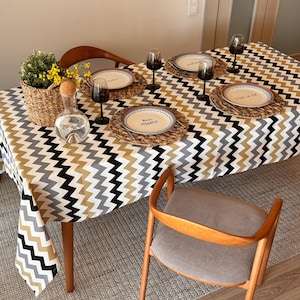 May include: A black, gray, and gold chevron patterned tablecloth with a woven placemat and a white plate with the text "Bon Appetit" on it. There are four wine glasses and a decanter on the table.