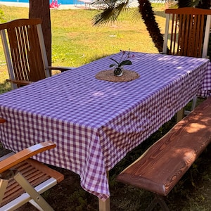 May include: A purple and white gingham tablecloth is laid out on a wooden table with two wooden chairs and a wooden bench. A small potted plant with pink flowers sits in the centre of the table.