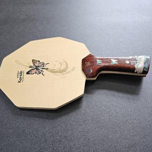 May include: A wooden ping pong paddle with a light-colored octagonal face. The face features a butterfly illustration and the text "Hello Korean". The handle is dark wood with decorative inlays.