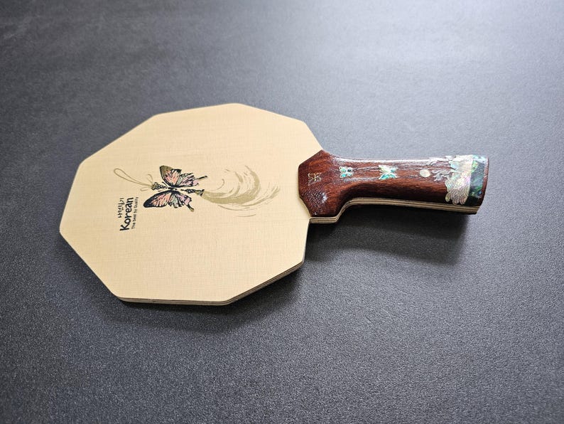 May include: A wooden ping pong paddle with a light-colored octagonal face. The face features a butterfly illustration and the text "Hello Korean". The handle is dark wood with decorative inlays.