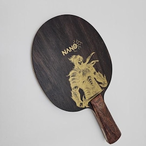 K&ouml;nnte beinhalten: Ein Tischtennisschl&auml;ger aus dunklem Holz mit einer goldenen Illustration eines Kriegers. Der Griff ist braun mit einem Muster. Das Wort "NANO" ist in Gold auf dem Schl&auml;ger aufgedruckt.