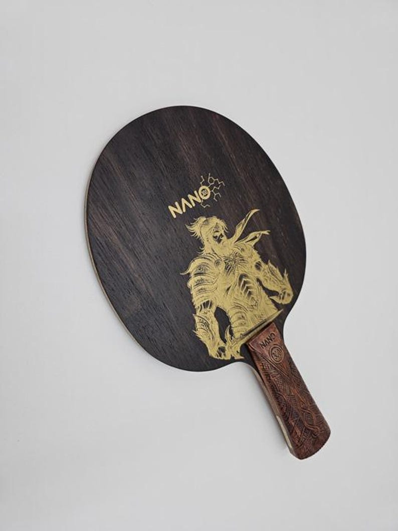 K&ouml;nnte beinhalten: Ein Tischtennisschl&auml;ger aus dunklem Holz mit einer goldenen Illustration eines Kriegers. Der Griff ist braun mit einem Muster. Das Wort "NANO" ist in Gold auf dem Schl&auml;ger aufgedruckt.