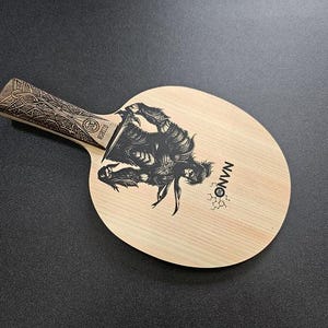 May include: A wooden table tennis paddle with a black and white graphic of a warrior on the surface. The paddle has a black handle with an intricate design and the word "NANO" printed on the surface.