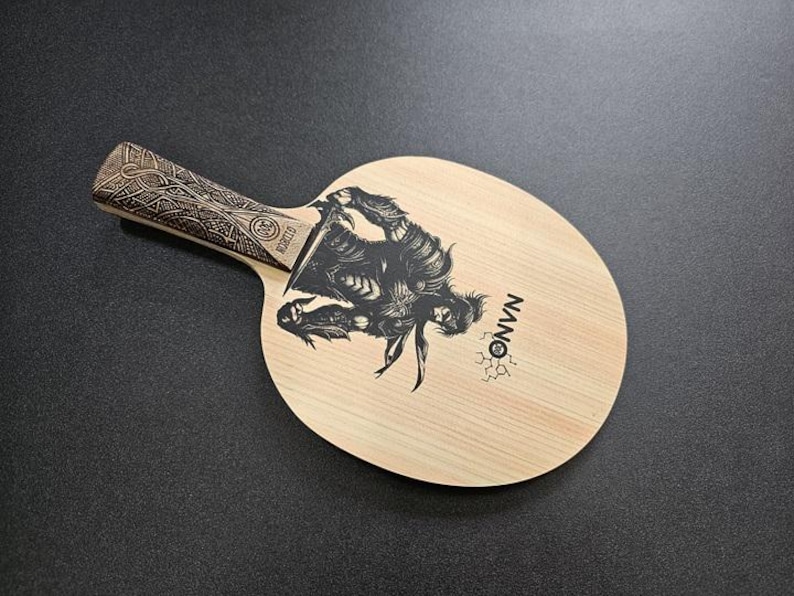May include: A wooden table tennis paddle with a black and white graphic of a warrior on the surface. The paddle has a black handle with an intricate design and the word "NANO" printed on the surface.