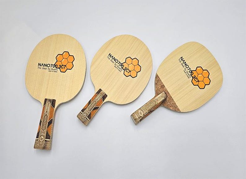 May include: Three ping pong paddles with light wood faces and patterned handles. Each paddle features a honeycomb graphic and the text "NANO700.3C7 The Best by Quality Made in Korea."