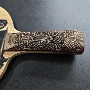 May include: A wooden table tennis paddle with a black and white graphic of a dragon on the front. The handle is made of wood and has a detailed carved design.