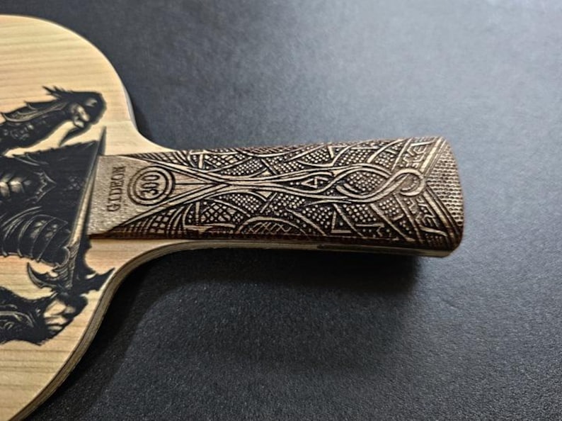 May include: A wooden table tennis paddle with a black and white graphic of a dragon on the front. The handle is made of wood and has a detailed carved design.