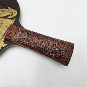 K&ouml;nnte beinhalten: Ein Tischtennisschl&auml;ger aus Holz mit einem goldenen Drachendesign. Der braune Griff hat ein aufwendiges Schnitzmuster. Auf dem Griff steht der Text "NANO (300)".