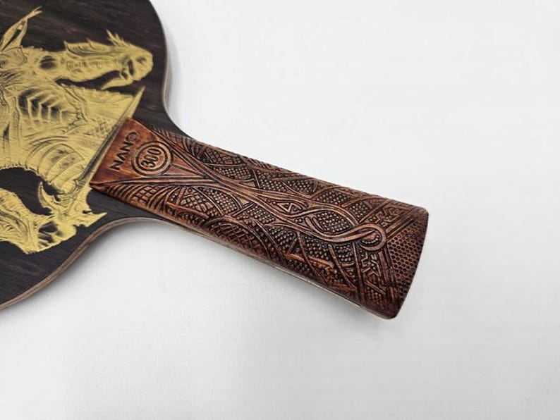K&ouml;nnte beinhalten: Ein Tischtennisschl&auml;ger aus Holz mit einem goldenen Drachendesign. Der braune Griff hat ein aufwendiges Schnitzmuster. Auf dem Griff steht der Text "NANO (300)".