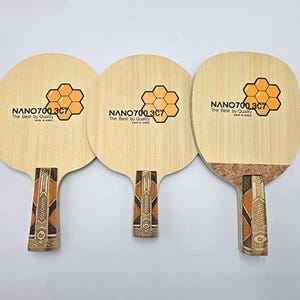 May include: Three light-colored wooden table tennis blades featuring a honeycomb graphic and the text NANO700 3C7 The Best by Quality Made in Korea. Each blade has a patterned handle with geometric brown and black accents and a cork grip.