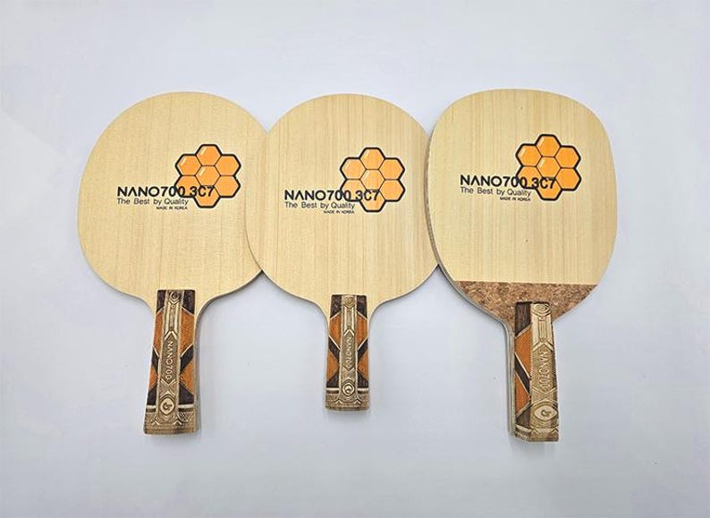 May include: Three light-colored wooden table tennis blades featuring a honeycomb graphic and the text NANO700 3C7 The Best by Quality Made in Korea. Each blade has a patterned handle with geometric brown and black accents and a cork grip.