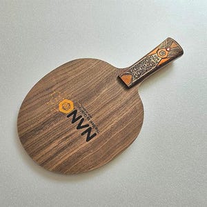 Puede incluir: Una raqueta de ping pong de madera con una hoja marr&oacute;n oscuro y un mango decorativo. El mango presenta un dise&ntilde;o naranja y marr&oacute;n. La raqueta tiene el texto "NVN The Best By Quality" impreso en la hoja.