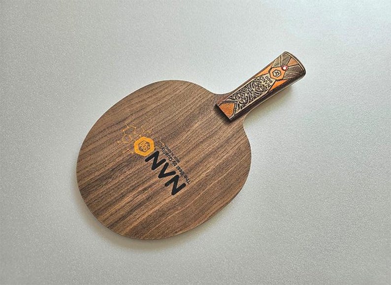 Puede incluir: Una raqueta de ping pong de madera con una hoja marr&oacute;n oscuro y un mango decorativo. El mango presenta un dise&ntilde;o naranja y marr&oacute;n. La raqueta tiene el texto "NVN The Best By Quality" impreso en la hoja.