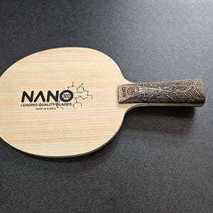 May include: A wooden table tennis paddle with a black and white logo that says "NANO 300 Leading Quality Blades Made in Korea". The handle is brown with a decorative pattern.
