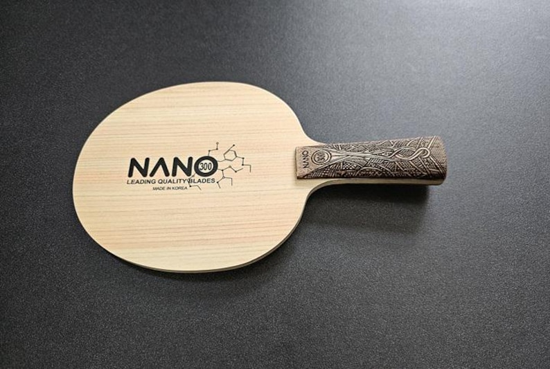 May include: A wooden table tennis paddle with a black and white logo that says "NANO 300 Leading Quality Blades Made in Korea". The handle is brown with a decorative pattern.