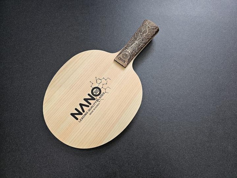 May include: A wooden table tennis paddle with a brown handle and a natural wood blade. The blade has the text "NANO 300 LEADING QUALITY BLADES MADE IN CHINA" printed on it.
