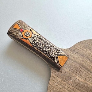 Puede incluir: Primer plano de una raqueta de ping pong de madera. El mango presenta detalles en naranja y marr&oacute;n con el texto "NANO 900" y un dise&ntilde;o decorativo. Una peque&ntilde;a gema roja est&aacute; engastada en el mango.