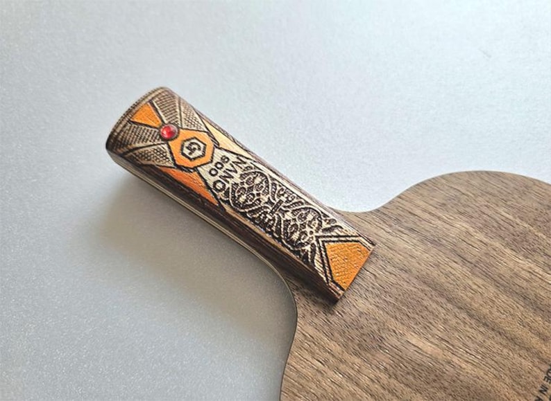Puede incluir: Primer plano de una raqueta de ping pong de madera. El mango presenta detalles en naranja y marr&oacute;n con el texto "NANO 900" y un dise&ntilde;o decorativo. Una peque&ntilde;a gema roja est&aacute; engastada en el mango.