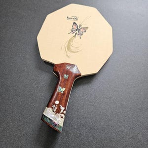 May include: A wooden ping pong paddle with a butterfly design and the text "Korean". The paddle has a brown handle with inlaid mother-of-pearl and butterfly accents. The paddle's head is an octagon shape.