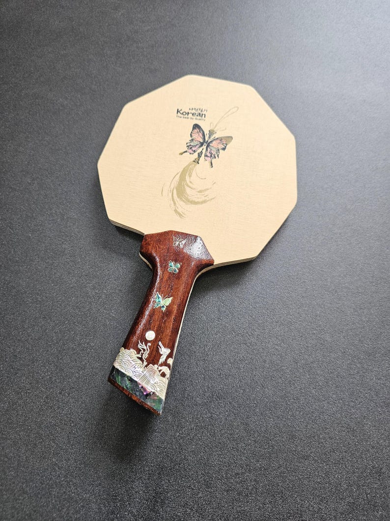 May include: A wooden ping pong paddle with a butterfly design and the text "Korean". The paddle has a brown handle with inlaid mother-of-pearl and butterfly accents. The paddle's head is an octagon shape.