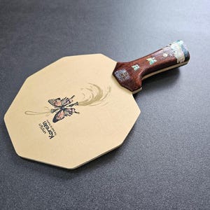 May include: A ping pong paddle with a light brown octagonal face and a dark brown handle. The paddle face features a butterfly illustration with the text "Korean" and a gold swirl design. The handle has decorative inlays.
