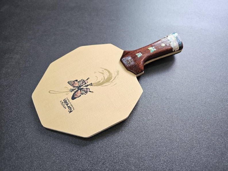 May include: A ping pong paddle with a light brown octagonal face and a dark brown handle. The paddle face features a butterfly illustration with the text "Korean" and a gold swirl design. The handle has decorative inlays.