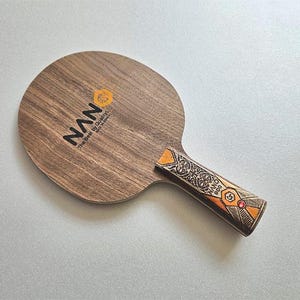 Puede incluir: Una raqueta de ping-pong de madera con la marca "NAN" impresa en negro. El mango presenta un dise&ntilde;o ornamentado con detalles en naranja y rojo. La raqueta es de madera y tiene forma redonda.