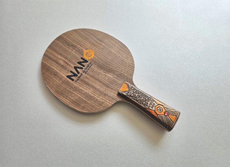 Puede incluir: Una raqueta de ping-pong de madera con la marca "NAN" impresa en negro. El mango presenta un dise&ntilde;o ornamentado con detalles en naranja y rojo. La raqueta es de madera y tiene forma redonda.