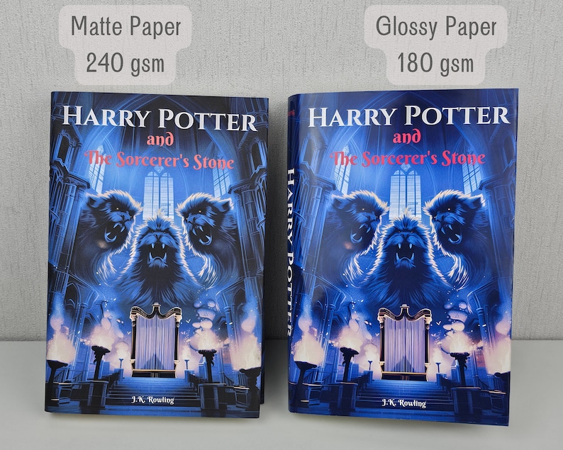 K&ouml;nnte beinhalten: Zwei Exemplare des Buches Harry Potter und der Stein der Weisen, eines mit einem matten Papierumschlag und eines mit einem gl&auml;nzenden Papierumschlag. Die Buchumschl&auml;ge zeigen eine blaue und schwarze Illustration von drei Greifen, die einen Thron bewachen.