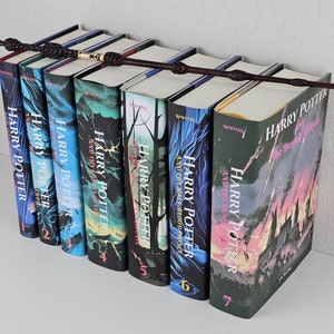K&ouml;nnte beinhalten: Ein Set aus sieben Harry-Potter-B&uuml;chern mit Illustrationen aus der Serie auf den Covern. Die B&uuml;cher sind in der Reihenfolge angeordnet, mit dem ersten Buch links und dem siebten Buch rechts. Ein schwarzer Zauberstab liegt auf den B&uuml;chern.