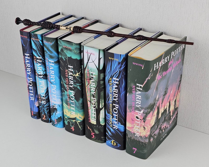 K&ouml;nnte beinhalten: Ein Set aus sieben Harry-Potter-B&uuml;chern mit Illustrationen aus der Serie auf den Covern. Die B&uuml;cher sind in der Reihenfolge angeordnet, mit dem ersten Buch links und dem siebten Buch rechts. Ein schwarzer Zauberstab liegt auf den B&uuml;chern.