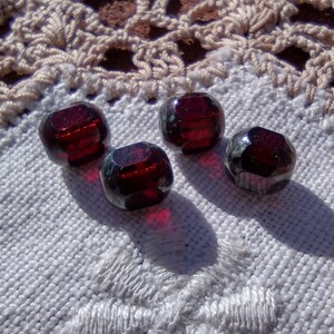 May include: Four dark red glass beads with a square shape and a slight green tint. The beads are on a white crocheted surface.