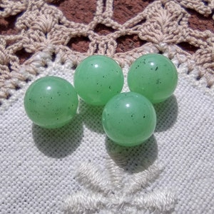 May include: Four translucent, light green glass marbles with dark speckles are arranged on a white embroidered surface. The marbles are round and smooth, casting shadows. A white, embroidered snowflake design is visible.