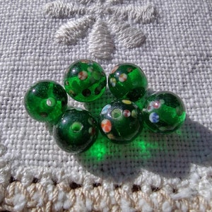 May include: Six green glass beads with white and multi-colored flecks. The beads are arranged in a circle on a white crocheted doily.