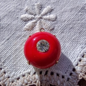 May include: A red round pendant with a clear rhinestone in the center. The pendant is on a white crocheted doily.