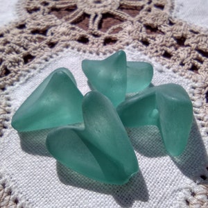 May include: Four sea glass pieces in shades of teal and aqua, arranged on a white crocheted doily.