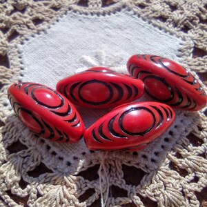 May include: Four red oval-shaped beads with black swirling designs. The beads are on a white crocheted doily.