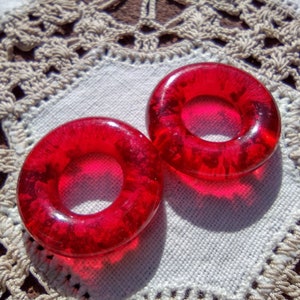 May include: Two red glass rings with a shiny, textured surface. The rings are on a white crocheted surface.