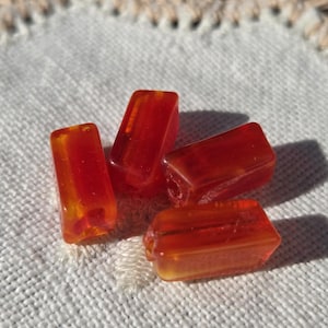 May include: Four translucent, rectangular, amber-colored beads are arranged on a white, textured surface. The beads have a polished appearance, with visible internal structures and variations in color. The background includes a crocheted lace edge.