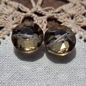 May include: Two faceted, round, amber-colored beads. Each bead has a central hole and a multifaceted surface that catches the light. The beads are set against a white fabric background.