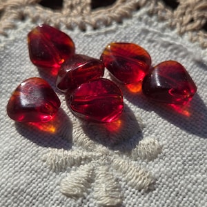 May include: A collection of seven translucent, faceted beads in shades of red and amber. The beads are irregularly shaped and appear to be made of glass or a similar material. They are arranged on a white, textured surface with a floral embroidery.