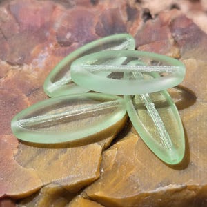 May include: Four translucent, light green oval-shaped beads with a textured interior. The beads are arranged on a rough, multi-colored stone surface, showcasing shades of brown, tan, and reddish-purple. These beads are suitable for jewelry making.