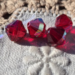 May include: Four faceted, deep red crystal beads are arranged on a white embroidered surface. The beads have a sparkling, iridescent quality, reflecting light. The background features a beige, open-weave fabric.
