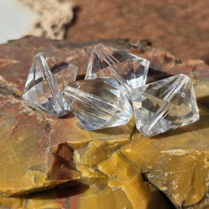 May include: Close-up of five clear, faceted crystal beads. The beads are diamond-shaped and reflect light, creating a sparkling effect. They rest on a multi-colored, textured stone surface, with a blurred background.